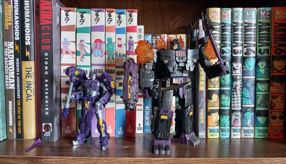 Two Transformers toys on robot form on a bookshelf with mainly Katsuhiro Otomo comics with a smattering of Moebius comics in background: 2 of the 13 original Primes, Solus Prime and Megatronus 