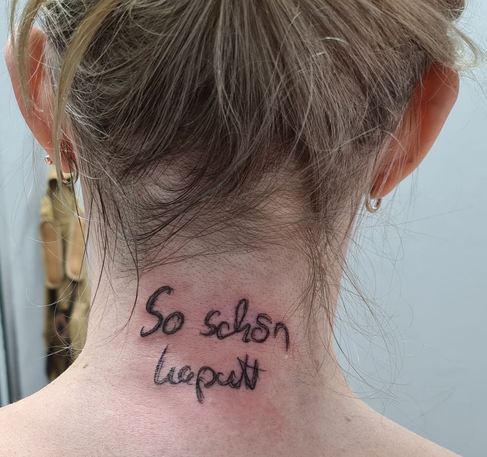 Foto meines Nackens, in dem frisch tätowiert die Worte "So schön kaputt" in meiner geschwungenen Handschrifr stehen. Die Haut unter dem Tattoo ist gerötet. Meine blonden Haare habe ich nach oben geknotet, damit der Nacken frei ist.