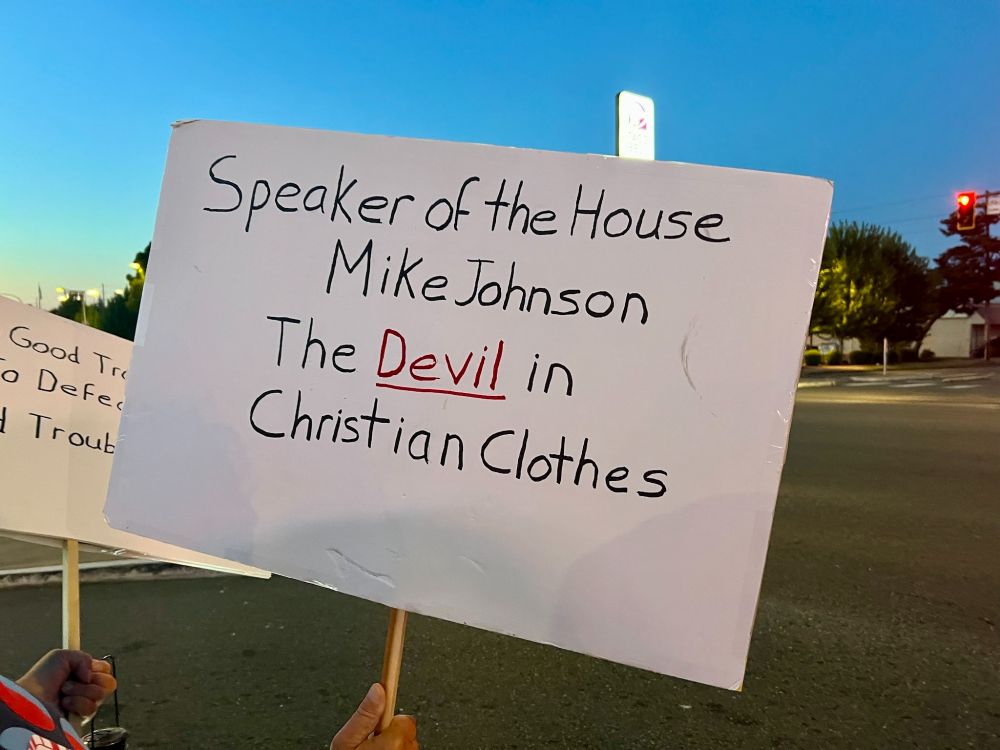 A protest sign reads: “Speaker of the House Mike Johnson — The Devil in Christian Clothes,” with the word “Devil” underlined and written in red, while the rest of the text is in black marker. The sign is held by a person standing near a street intersection during twilight, with a traffic light and trees visible in the background.