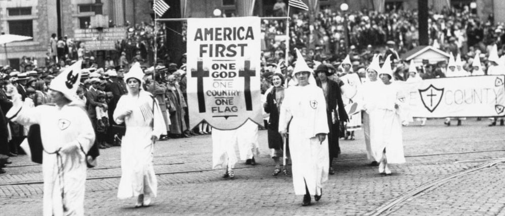 Black and white photo of a Ku Klux Klan parade in Binghamton, New York during the 1920s. Klan members, both men and women, are dressed in white robes and pointed hoods, marching down a cobblestone street. At the front, several participants carry an American flag and a large banner reading "AMERICA FIRST – ONE GOD, ONE COUNTRY, ONE FLAG" between two crosses. A crowd of onlookers stands behind police barriers. In the background, another Klan banner with a cross and shield symbol is partially visible.