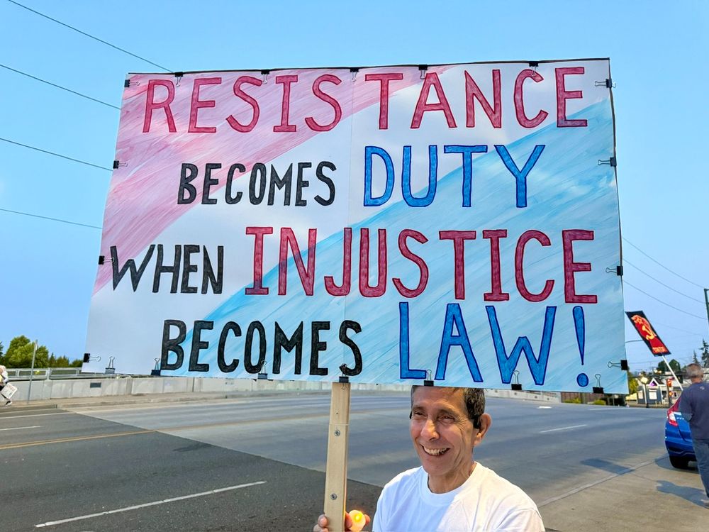 A protest sign held by a smiling man reads: “RESISTANCE becomes DUTY when INJUSTICE becomes LAW!” The words “RESISTANCE,” “INJUSTICE,” and “LAW!” are written in bold red, while “DUTY” and “LAW!” are in blue, and the remaining text in black. The background is diagonally shaded in light red on the left and light blue on the right, giving the sign a split-tone look. The scene takes place on a city street at dusk, with cars, other protesters, and street signs visible in the background.
