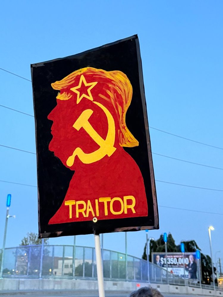 A protest sign shows the red silhouette of former President Donald Trump's profile, with his distinctive yellow-orange hair. His face is overlaid with a yellow hammer and sickle, symbolizing communism, and a red star above his forehead. Below the image, the word "TRAITOR" is written in bold yellow letters. The sign is held up outdoors during twilight, with power lines and buildings faintly visible in the background.