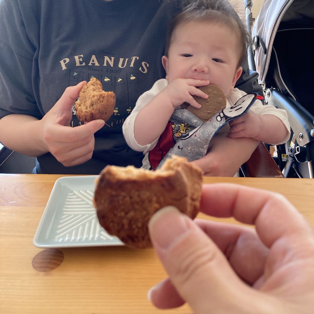 スコーンを手にする筆者の手（食卓手前）と、同じく妻（食卓向こう）と、オモチャのクッキー（歯固め用）を口にする子ども（妻の膝の上）