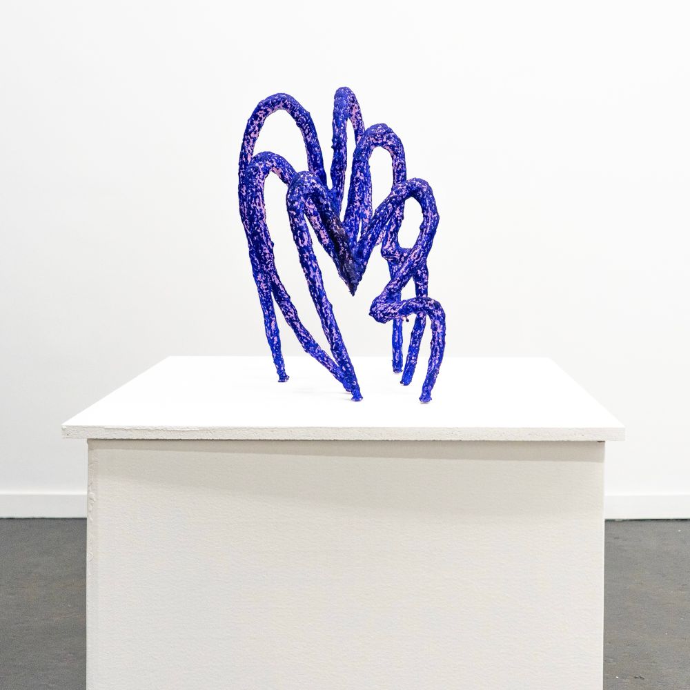 A bright blue and lavender sculpture with 6 limbs and arched spine creating an abstracted heart shape form, on a white pedestal.