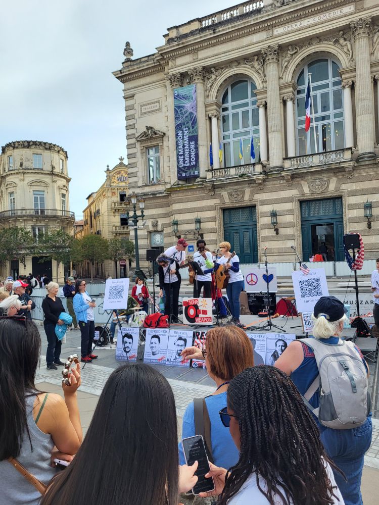 A group of musicians play onstage on front of an opera house. Posters in front of the stage call for solidarity with activists and immigrants targeted by the regime. 