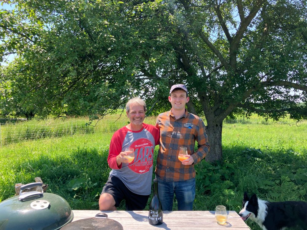 Sam and I standing together behind a picnic table and in front of an apple tree, each holding a glass of cider. A bottle on the table reads “2024 Wild - Trees 2 + 5.”