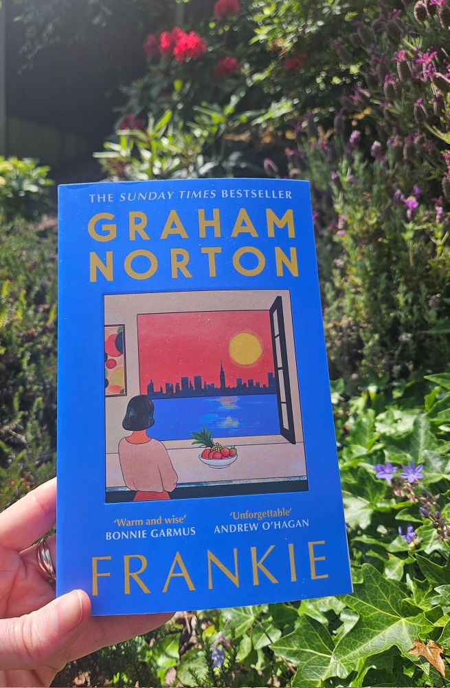 A hand holding a copy of Frankie, a book by Graham Norton, in front of foliage and flowers
