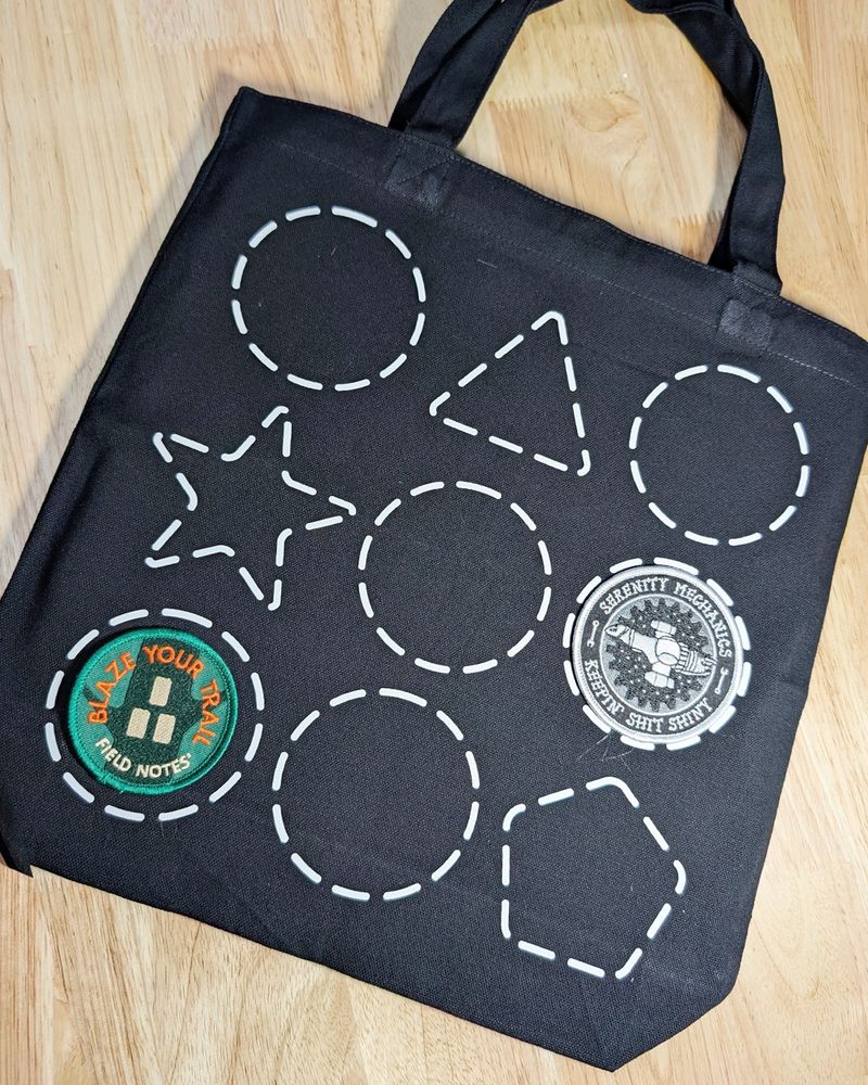 A black tote bag with a design of shapes outlined in dashed lines. There are 3 lines of shapes, each featuring two circles, and one unique shape including a triangle, a star, and a Pentagon. Two of the circles have patches centered in them.