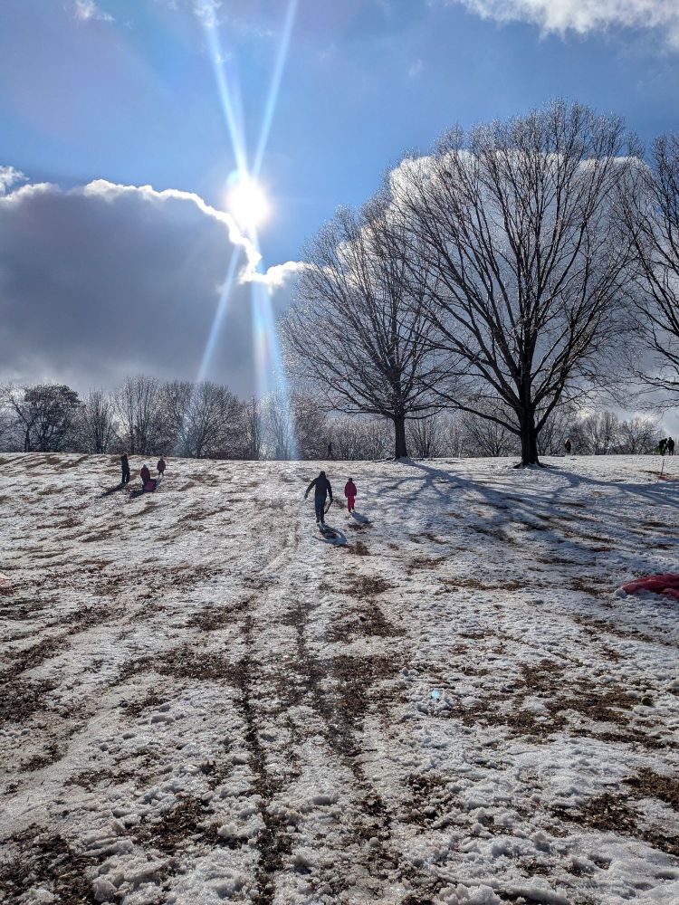Two people walk up a snowy hill while others prepare to sled down