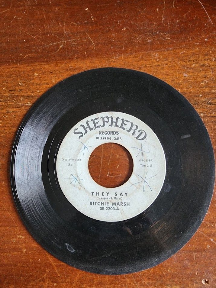 A 7-inch vinyl 45 rpm phonograph record laying on a wooden table. The label is off-white in color with grey text, including the logo for Shepherd Records, Hollywood, California; the title of the A-side, "They Say"; and the recording artist, Ritchie Marsh.
