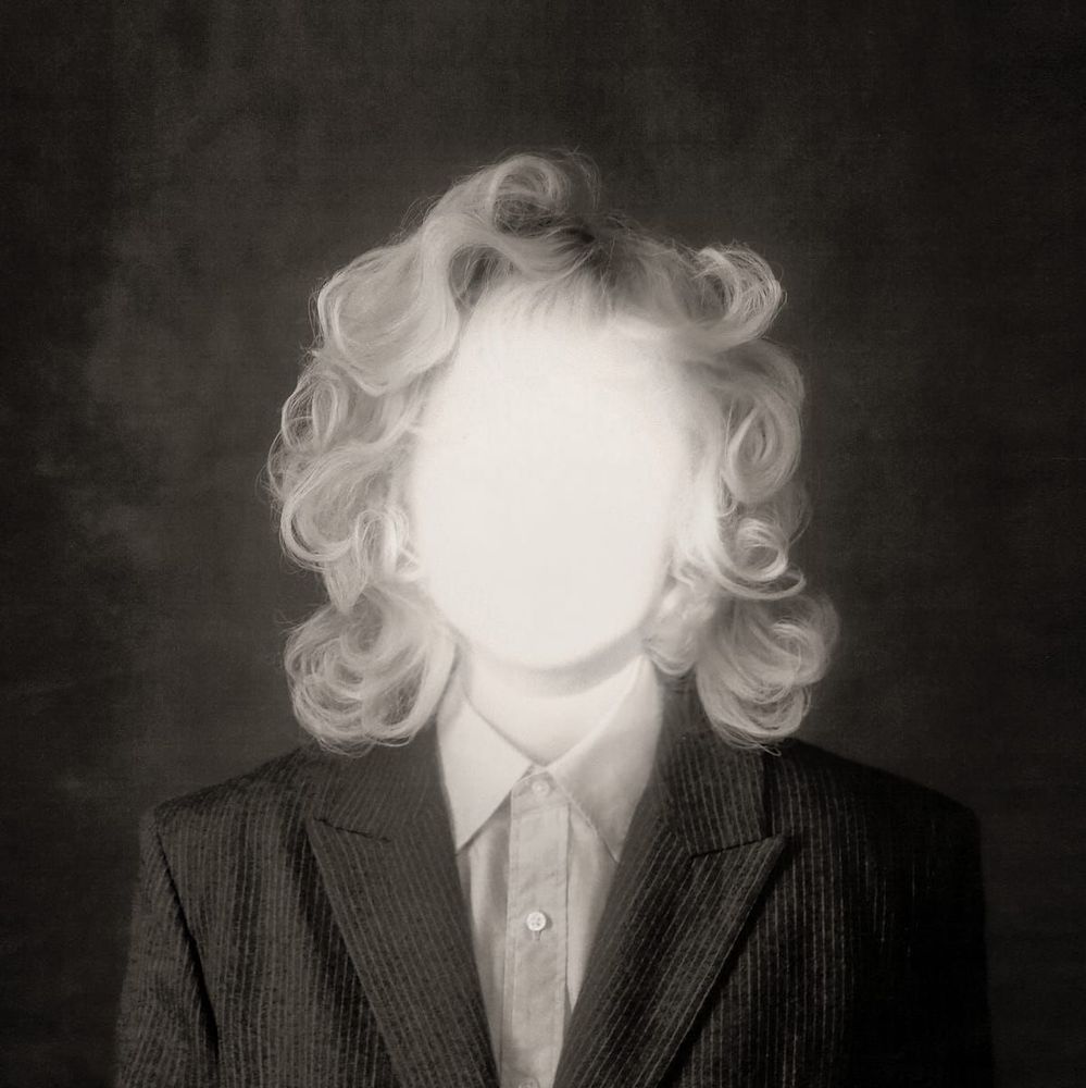 Black and white photo. A person wearing a suit and button shirt with blonde curls. There is a bright light shining on the face that washes the features out completely.