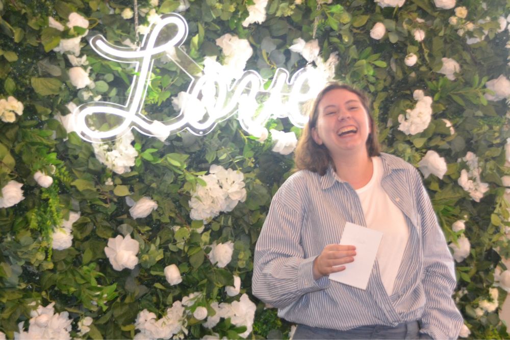 A picture of Catherine, in an open blue button down with a white shirt tucked into grey pants, laughing and standing in front of a green and white flower wall with a white neon sign that says “Love” in script.