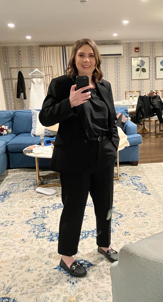 A full length mirror selfie of Catherine with one hand in her pocket. She is in an all black suit; the shirt a solid black satiny blouse tucked into ankle length pants. The black jacket is open, unbuttoned. Shoes are flat black loafers with a decorative silver buckle type thing.
She has some soft natural looking makeup on and her shoulder length hair has a bit of a subtle wave to it around her ears down.