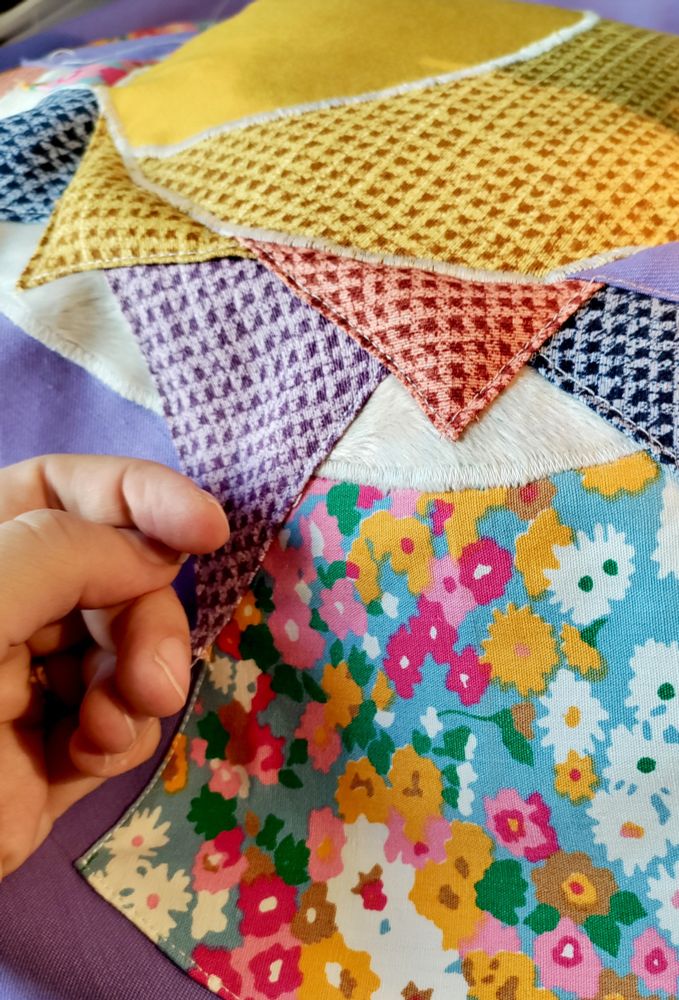 A hand next to the sewing project, which has a flowery fabric as well as purple and blue and red houndstooth and purple plain fabric and white fake minky.