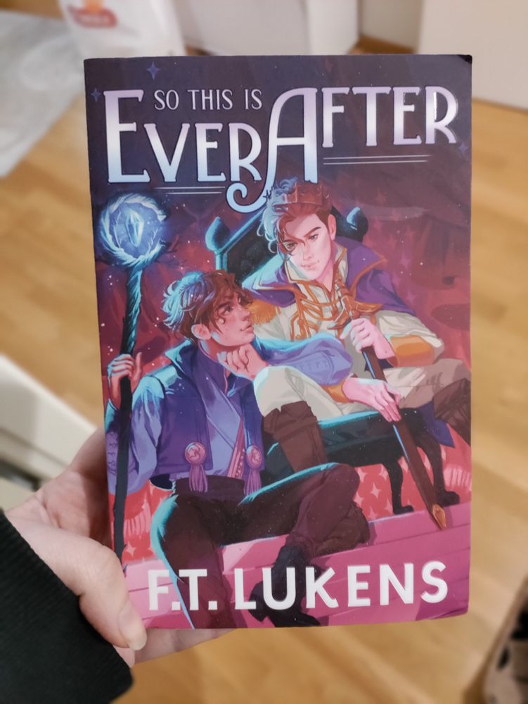 A hand holding So This Is Ever After by F.T. Lukens. The cover depicts two unnecessarily pretty teengers, one a king and the other a mage, gazing longingly at each other.