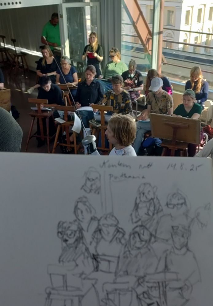 A photo of a life drawing audience observing a fencer in a drawing studio. In the foreground, there's a sketchpad with a quick sketch of the audience.