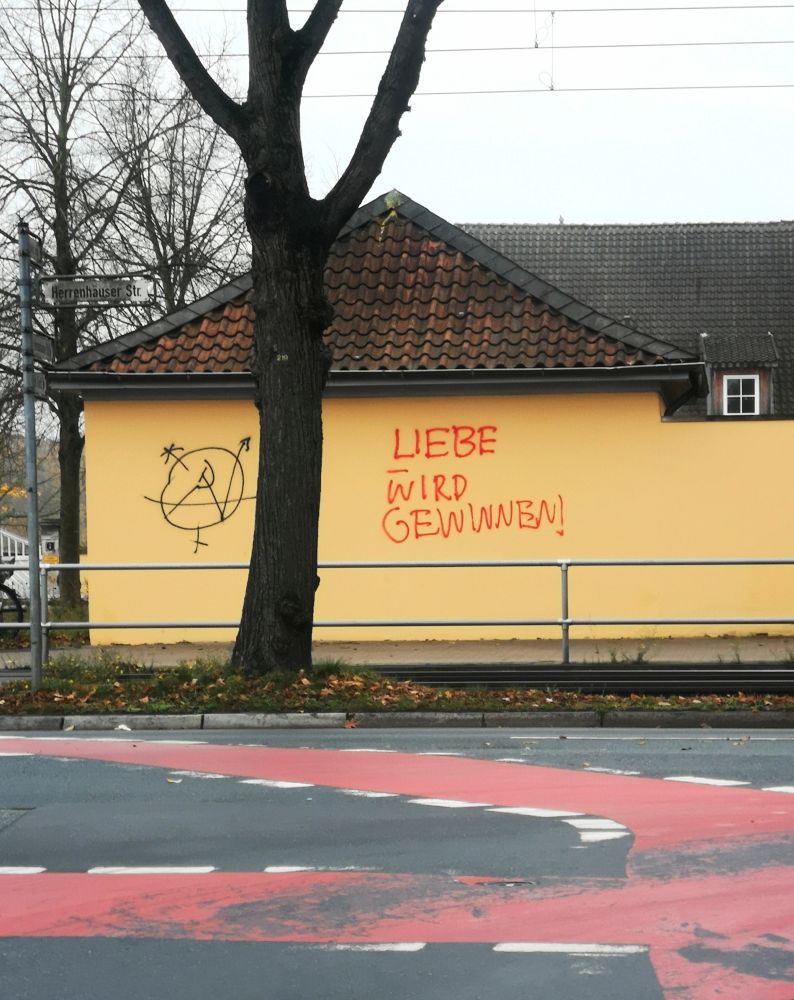 Hinter einer Straße, auf der ein Fahrradweg rot markiert ist, steht ein Haus mit einem neuen gelblichen Anstrich. Darauf ist in rot gesprüht: Liebe wird gewinnen! Links daneben noch ein seltsam kruckeliges Zeichen, ein Kreis mit einem Zickzack-Pfeil, ähnlich dem Hausbesetzer-Zeichen, evtl. mit einer Art Hammer & Sichel in der Mitte. Oder so. 