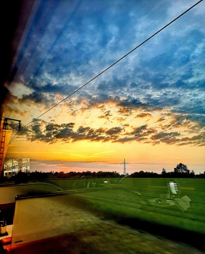 Schnappschuss aus dem Fenster des Speisewagens: Sonnenuntergang am Horizont, im Vordergrund grüne Felder, dunkelblauer Himmel mit dünner Wolkendecke. Einige Spiegelungen des Zugfensters, und man sieht auch das Spiegelbild einer Kaffeetasse. 