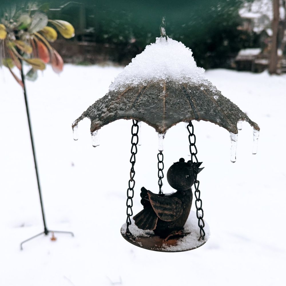 Yard art: a small metal bird sits on a platform attached to a metal umbrella with 3 chains. The umbrella and platform are covered in snow. Icicles hang from the points of the umbrella. Behind it is a snowy expanse of yard and a big metal flower.