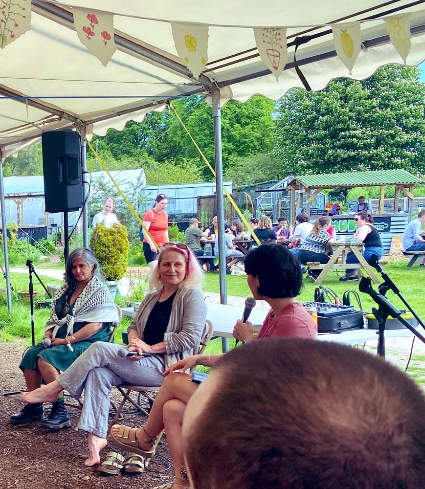 Two speakers and an interviewer with a microphone are sat underneath an awning. The floor is covered with woodchip, in the background is a community garden with lots of people walking or seated on table benches.
