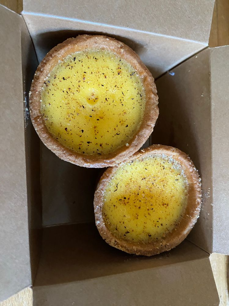 Overhead view of two custard tarts in a box, they are sprinkled with a nutmeg topping.