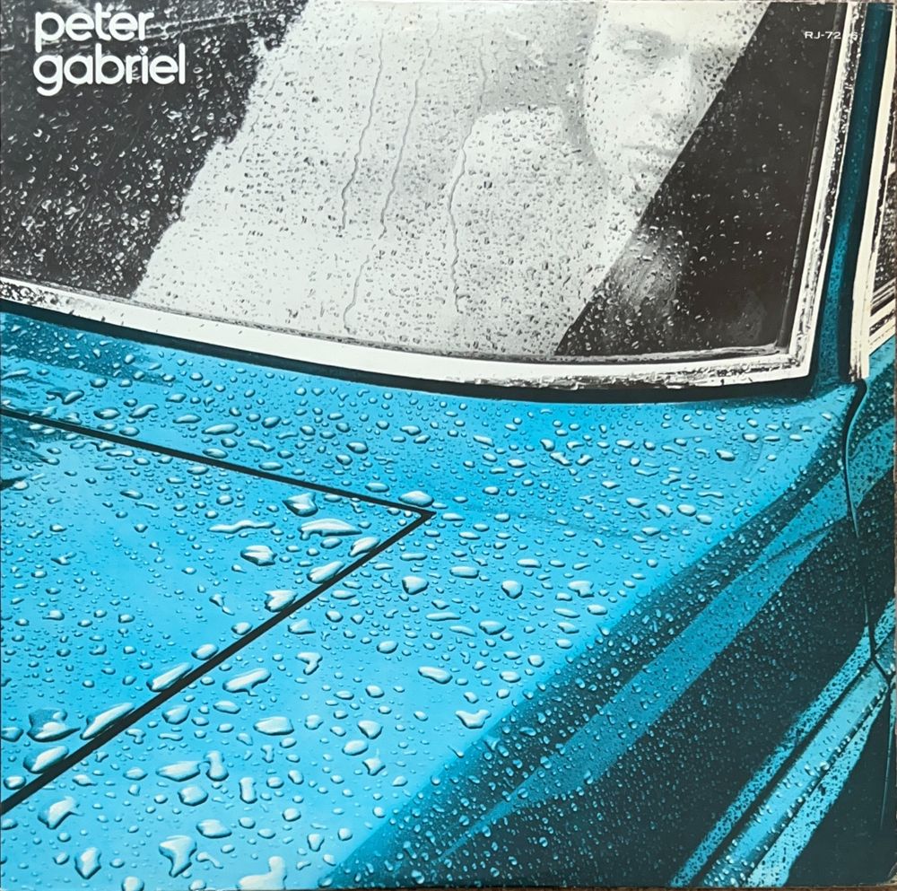 Album cover featuring a close-up of a blue Lancia Flavia car, with raindrops on the bonnet and the windscreen. A pale black and white face is visible in the passenger seat. It’s the artist Peter Gabriel, his eyes look odd, because he’s wearing mirrored contact lenses.