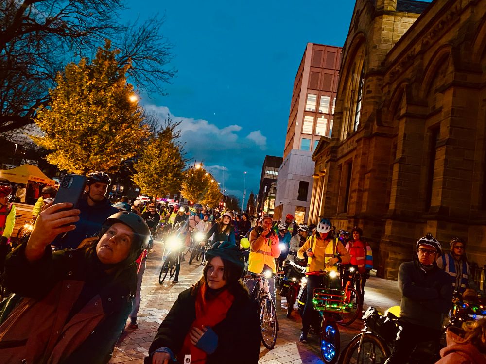 A group of people on bikes with lots of lights wait for a ride to set off as dusk descends. Trees and tall buildings can be seen either side of them.