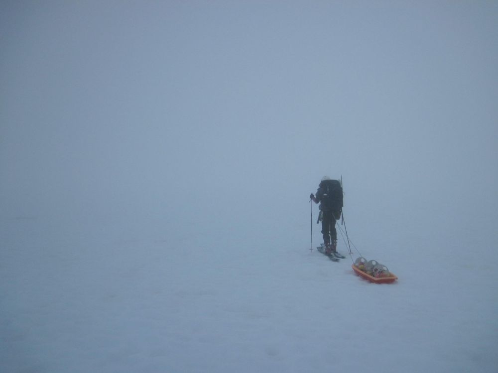Person on skis wearing a large backpack and pulling a sled over a snowy surface that melts into the thick gray fog above, with no horizon, sky, or other features visible.
