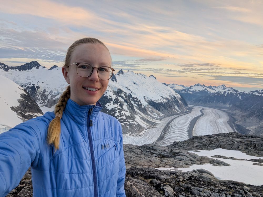 Selfie with a glacier in the background, surrounded by snow-covered mountains with clouds in the sky tinted yellow from sunset