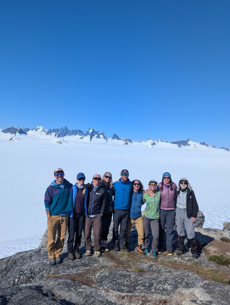 Nine people smiling in a row in front of a wide expanse of snow with dark peaks in the background