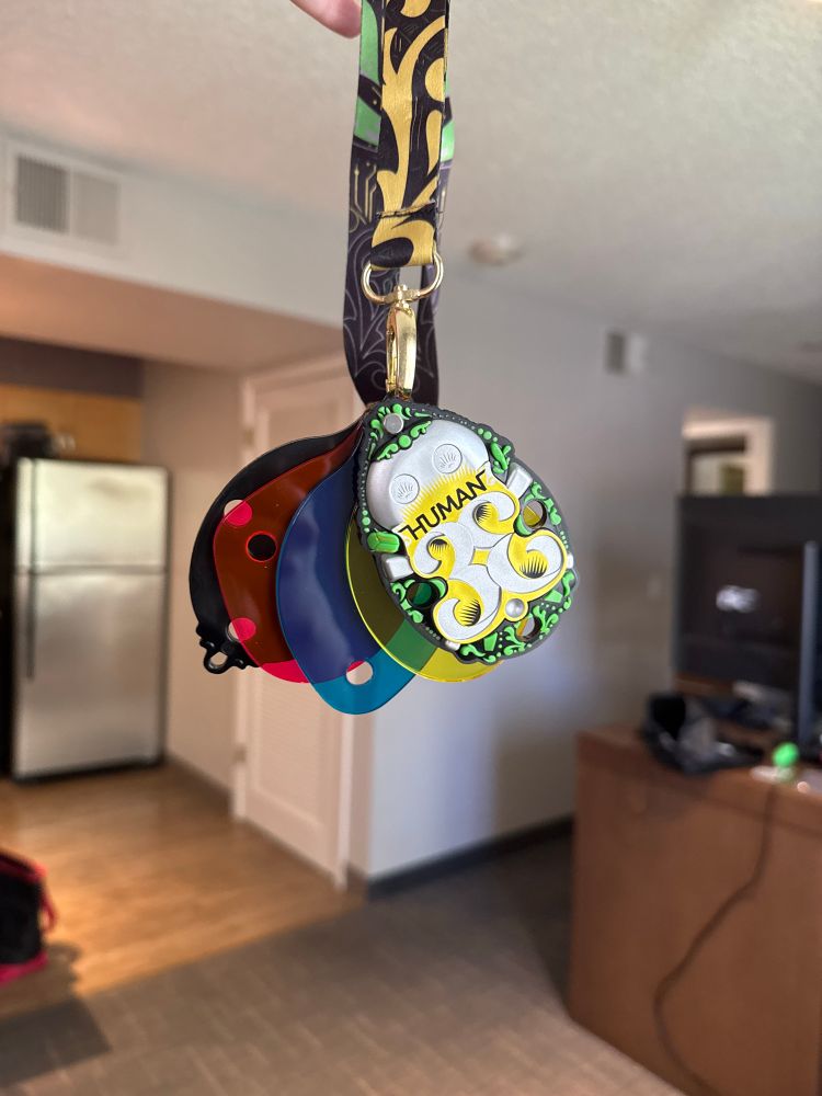 the DEF CON 33 HUMAN badge hanging by a lanyard in a hotel room. it’s 3 layers of clear colored acrylic (magenta, cyan, yellow) sandwiched between two black opaque acrylic layers which are the shell. the front is ornate with the DEF CON Jack skull and the number 33 where the right 3 is mirrored