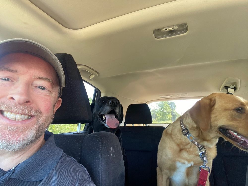 Old white guy with beard in passenger seat and a golden and black dog in the back seat of a vehicle. The man is smiling, the black dog has her tongue lolling out of her mouth. 