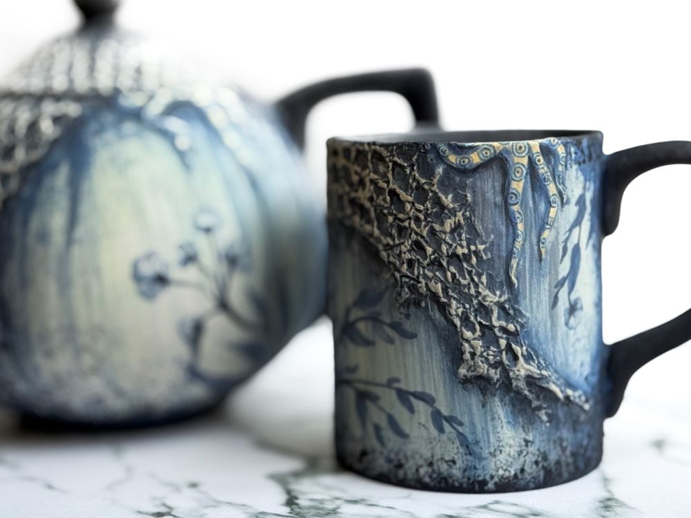 Teapot and mug featuring a weathered floral design with netting covering parts of each piece and a blue ring octopus crawling out of the top of both of them. Detail of mug