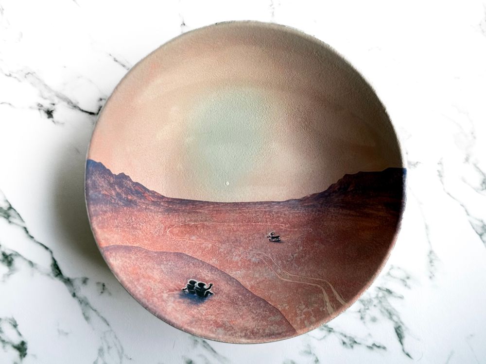 Bowl featuring two astronauts in the foreground sitting on Mars at sunset waving at a rover that travels inside a canyon followed by its own tracks and looks back at them. 