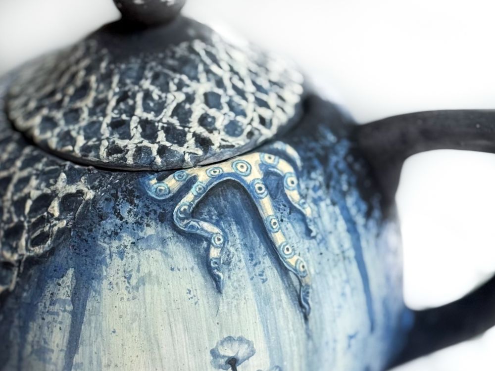 Teapot and mug featuring a weathered floral design with netting covering parts of each piece and a blue ring octopus crawling out of the top of both of them. Detail of blue ring octopus on teapot. 