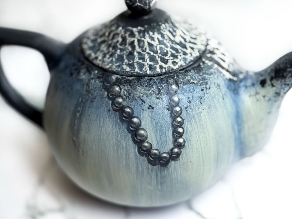 Teapot and mug featuring a weathered floral design with netting covering parts of each piece and a blue ring octopus crawling out of the top of both of them. Detail of back of teapot, showing pearls coming out from under lid. 