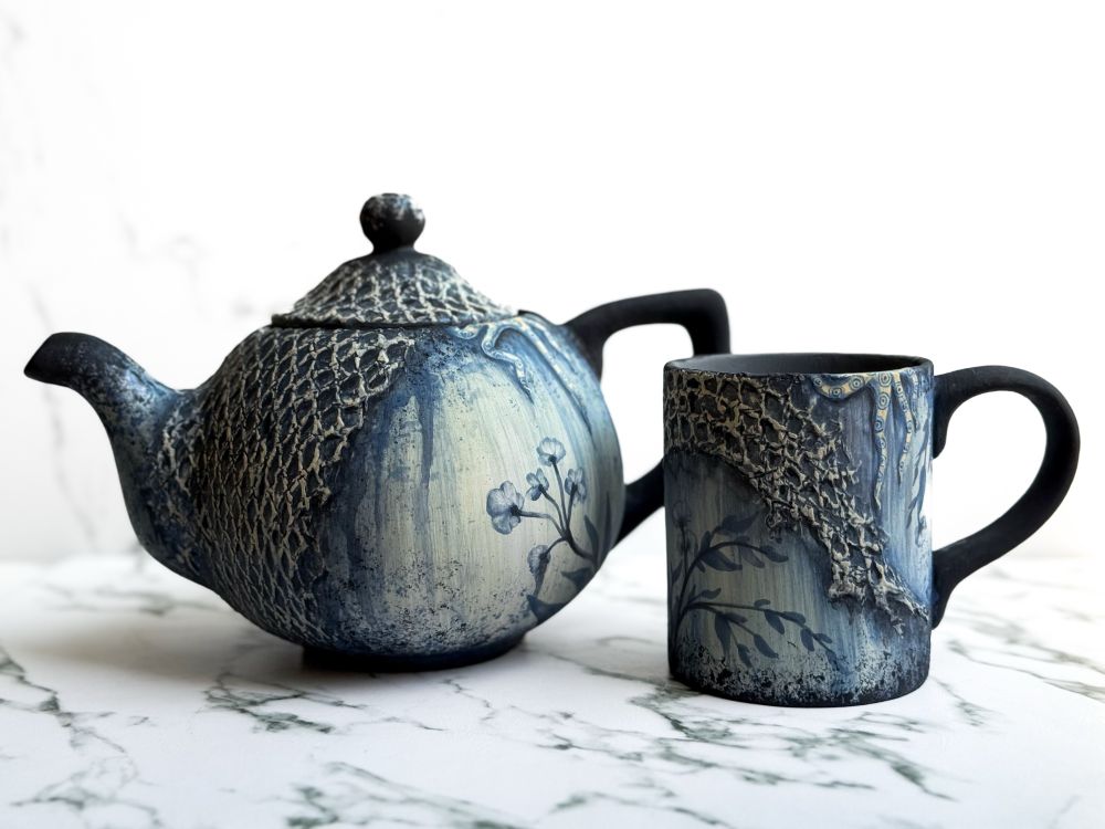 Teapot and mug featuring a weathered floral design with netting covering parts of each piece and a blue ring octopus crawling out of the top of both of them. 