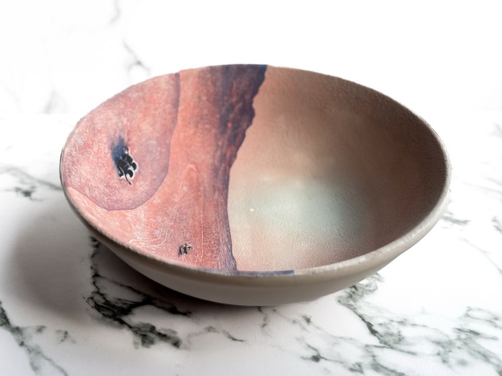 Bowl featuring two astronauts in the foreground sitting on Mars at sunset waving at a rover that travels inside a canyon followed by its own tracks and looks back at them. (Side)