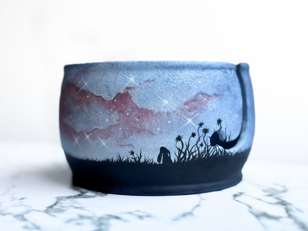 Yarn bowl featuring silhouette of floppy-eared rabbit in tall grass looking up at a red and pale purple nebula with stars shining around it. 