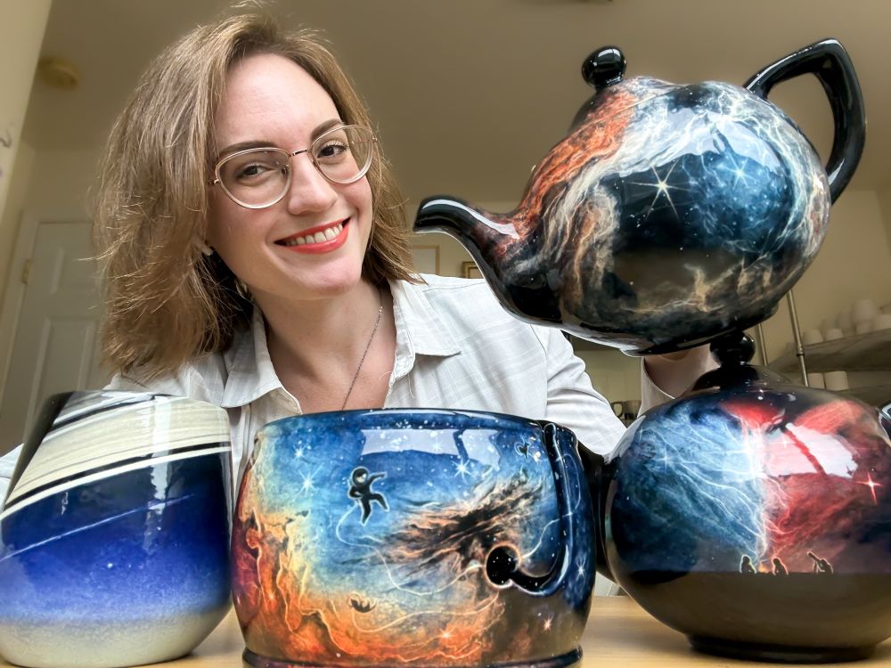 Me smiling holding a tarantula nebula teapot with a teapot, yarn bowl, and angled cup in front me featuring two different nebulae and the Cassini Pale Blue Dot. 