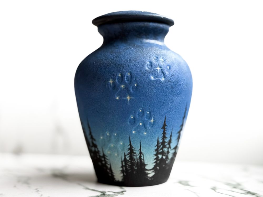 Urn with lid featuring silhouette of a dog looking up toward the Milky Way in the parting of a forest. Back showing paw print impressions in the sky highlighted with stars. 