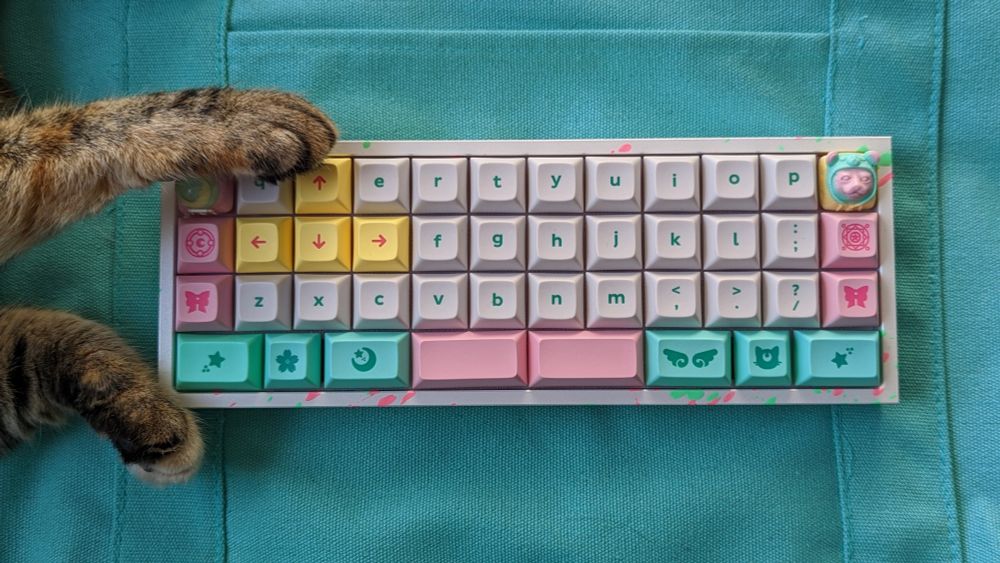 A white Gridiron 40% keyboard that has pink and green splashes and DSA Magic Girl keycaps. The keyboard is sitting on a teal tote bag and a tabby cat is reaching from out of frame and touching the left side of the keyboard with its paws