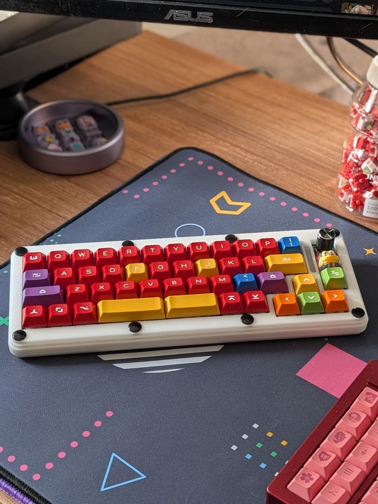 A white stacked acrylic Program Yoink keyboard sitting on a desk with colorful DSS Hand Work keycaps. The keyboard has a black rotary encoder as well as an artisan keycap from Rubrehose that matches the other keycaps.