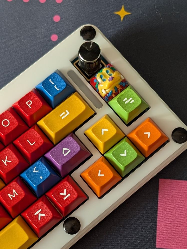 An angled shot of the right side of a white stacked acrylic Program Yoink keyboard sitting on a desk with colorful DSS Hand Work keycaps. The keyboard has a black rotary encoder as well as an artisan keycap from Rubrehose that matches the other keycaps.