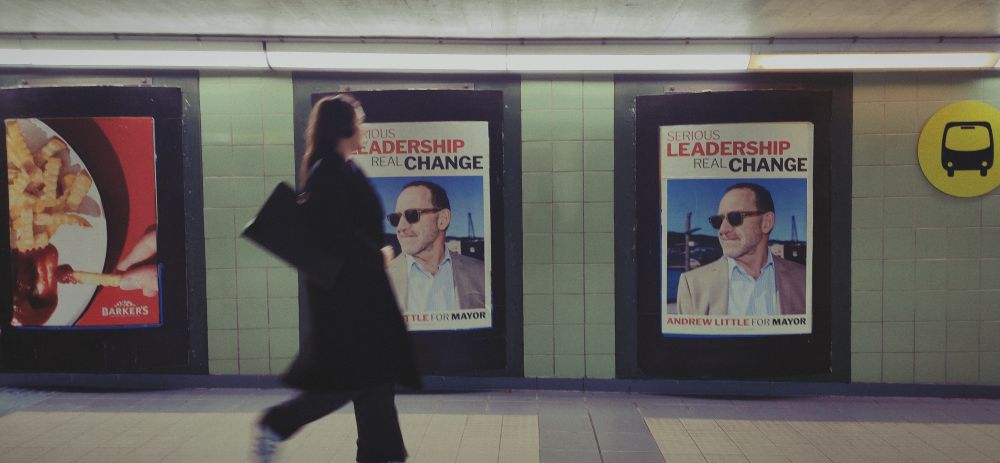 A commuter walks past two posters of Andrew Little in sunglasses looking like a capable Whyte guy who's relaxed but "capable" in the sunshine with the tag line "Serious Leadership, Real Change". Positioning himself in contrast to Mayor Whānau giving serious let a Whyte guy handle it love vibes. 