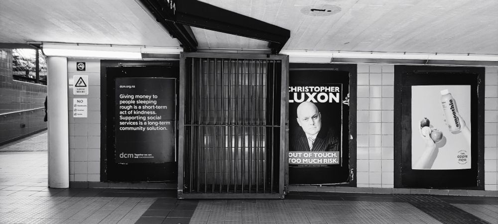 A black and white photo of the Wellington train station underground in 2023 with a poster saying Christopher Luxon too out of touch, too much risk. Next to a poster saying giving money to people sleeping rough is short term kindness - supporting community services is a little big term solution.