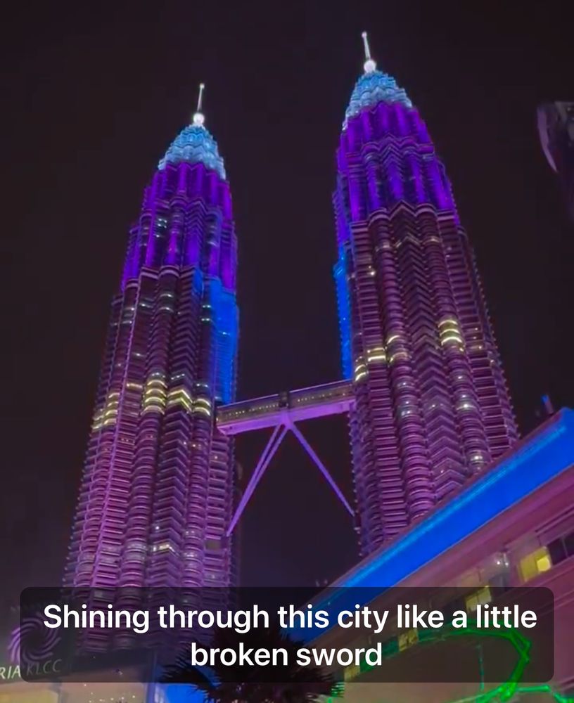 A screenshot of a BTS Dynamite lyric video showing two tall skyscrapers lit up in shades of purple with the caption “Shining through this city like a little broken sword” instead of the actual lyrics “Shining through the city with a little funk and soul” 