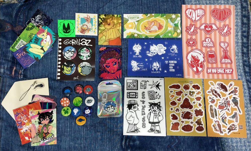 various colorful printed artist merch laid out on my blanket. a small pile of business cards, a small pile of swap cards, a few stickerpacks and button pins