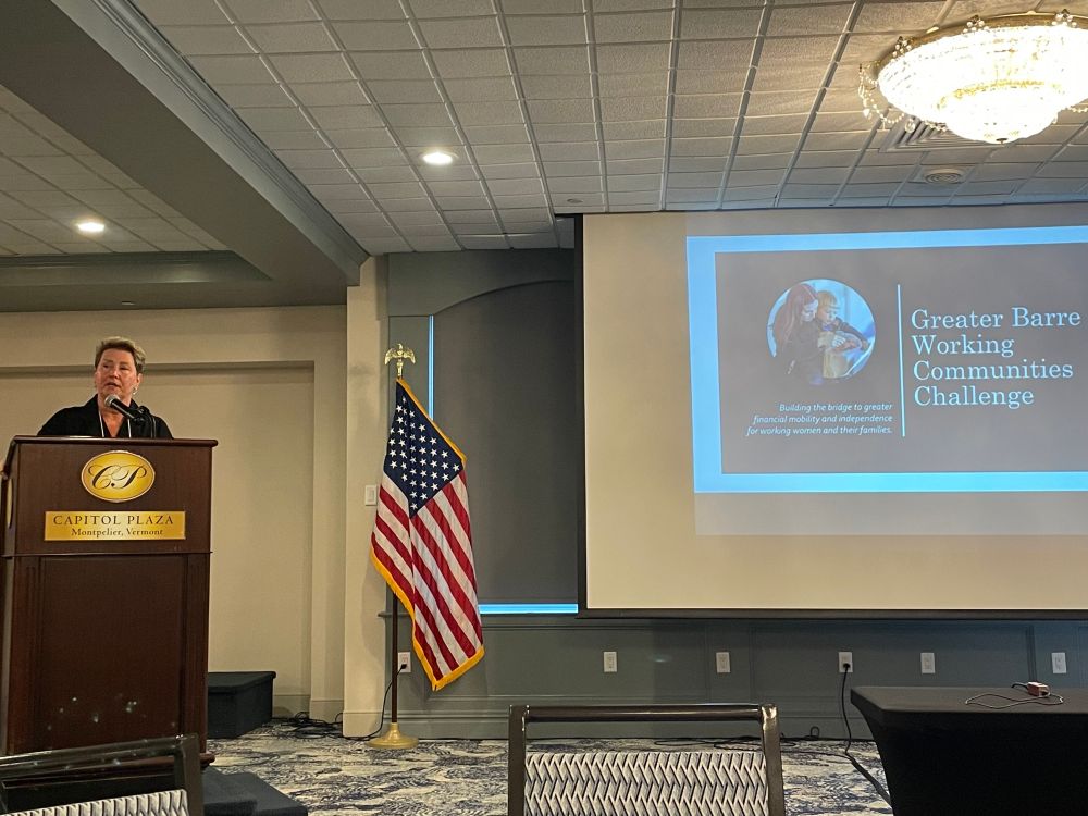 A photograph of a white woman standing at a podium speaking to an audience in front of a powerpoint that reads "Greater Barre Working Communities Challenge."