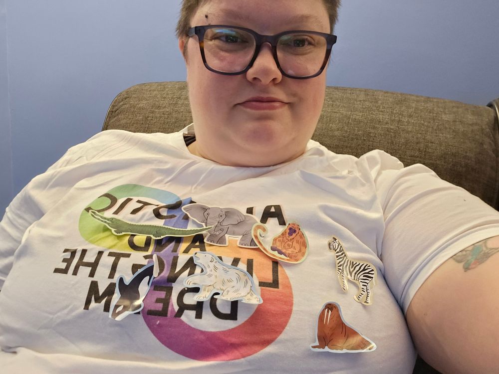 Me wearing a white Autistic and Living the Dream t shirt covered in animal stickers: a crocodile, elephant, walrus, zebra, monkey, polar bear, and a killer whale. All the classics.
